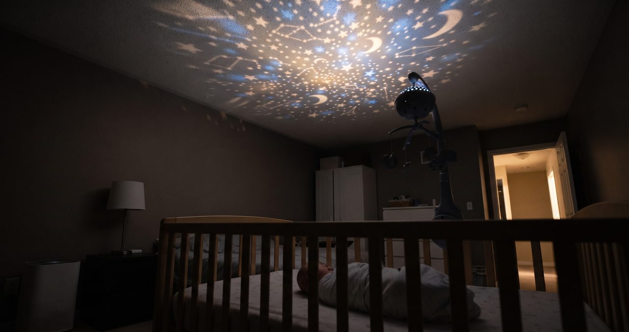 薄暗い寝室の天井に電動メリーが星空のような光の模様を投影している様子、ベビーベッドの柵がシルエットで見える引きのショット